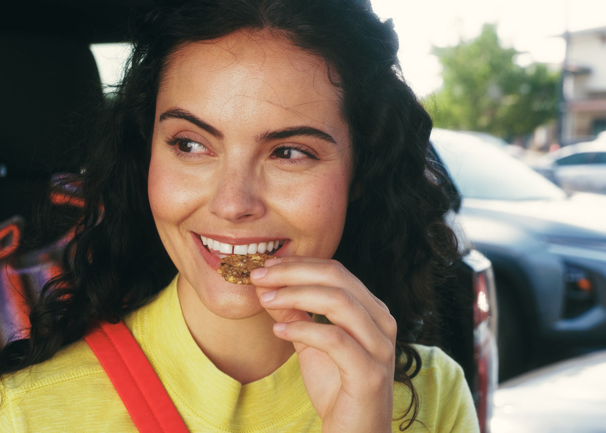 A smiling woman with dark curly hair wearing a yellow shirt and a red shoulder strap, standing in front of an open car door with parked cars in the background.
